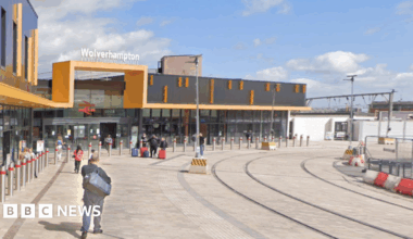 Passengers make their way to the train station in Wolverhampton. It has a yellow and black entrance with an old British Rail sign above the entrance and tram lines running past the front a few feet away.