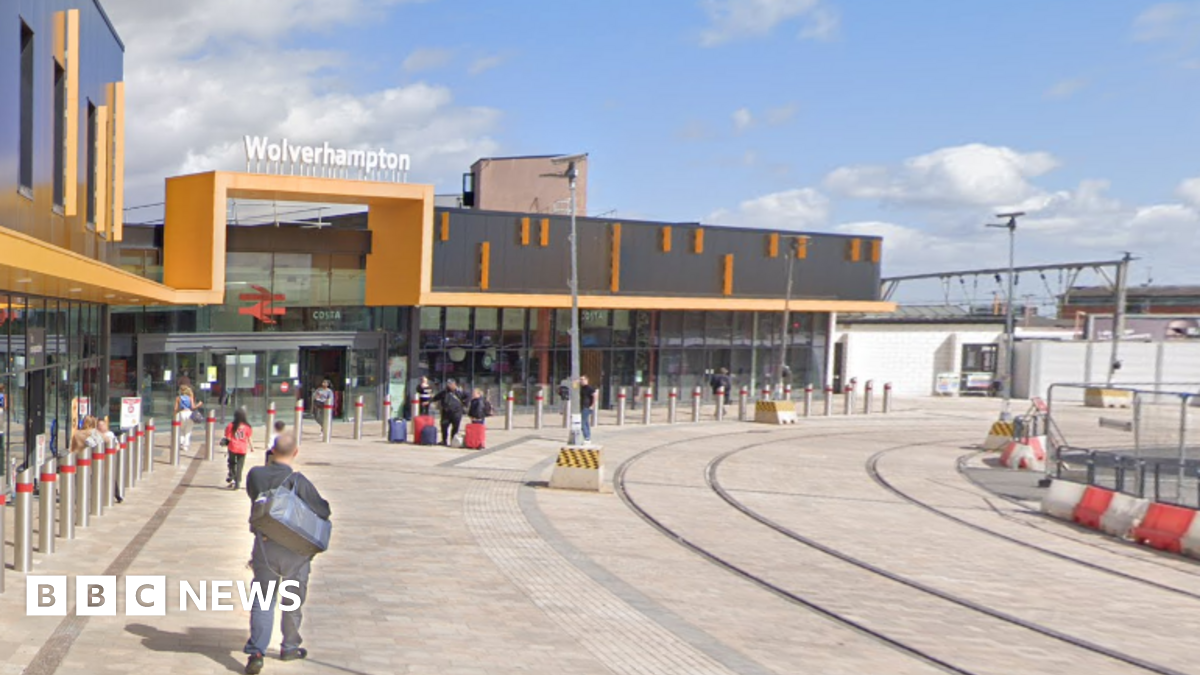 Passengers make their way to the train station in Wolverhampton. It has a yellow and black entrance with an old British Rail sign above the entrance and tram lines running past the front a few feet away.