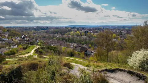 Getty Images Panoramic view of a suburban area with houses surrounded by trees and greenery. Dirt paths wind through grassy and bushy terrain in the foreground. The background features forested areas and distant hills under a partly cloudy sky.