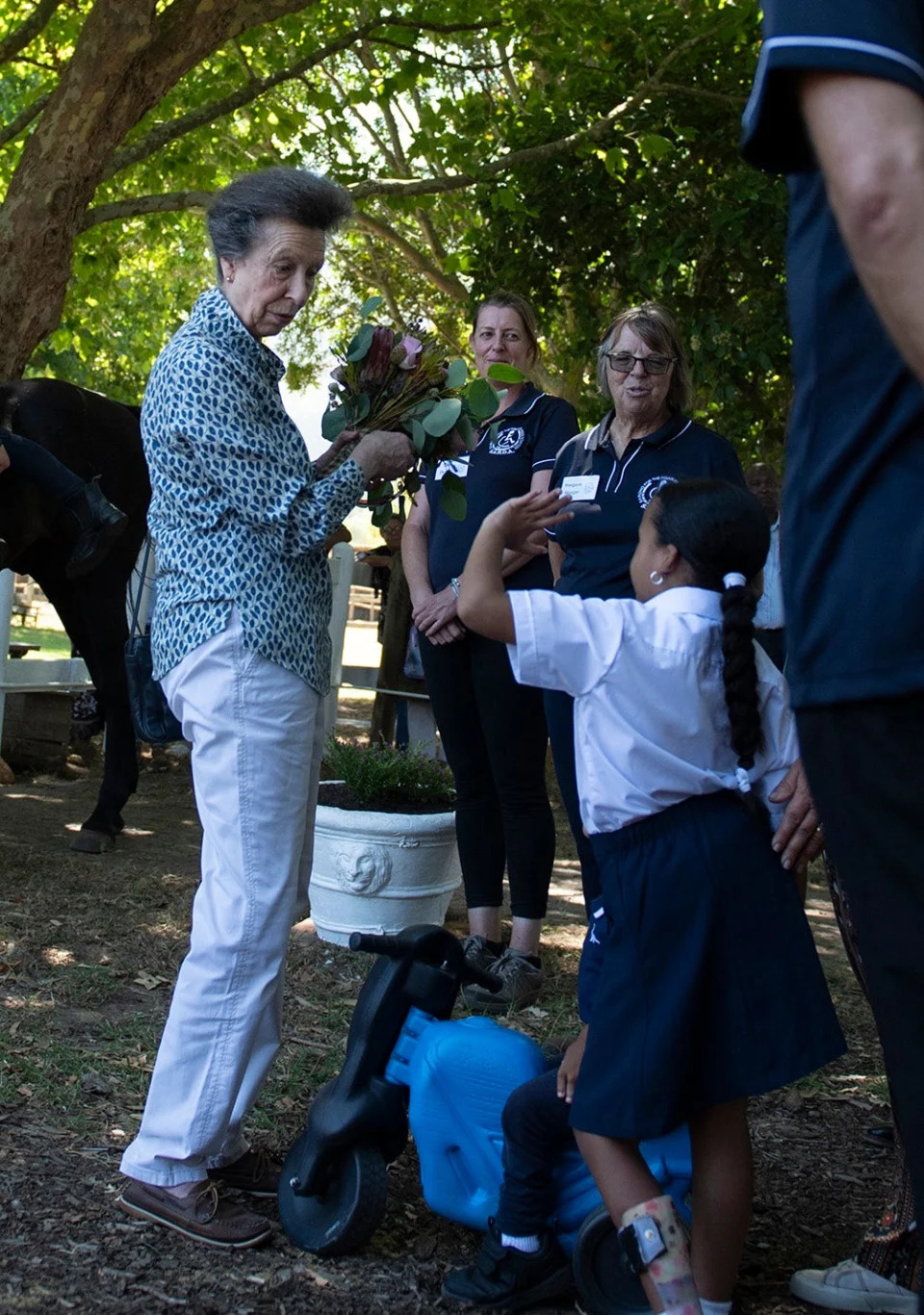 Princess Anne wearing a blue multiprinted blouse and white pants interacting with people outdoors.