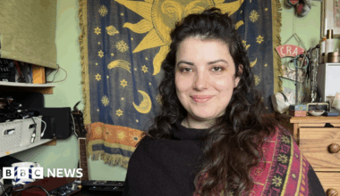 Lily poses for a photograph next to her recording and mixing equipment in her bedroom in Leominster. Behind her are shelves of electronic equipment, a tapestry with the Sun, moons and stars, and a guitar.