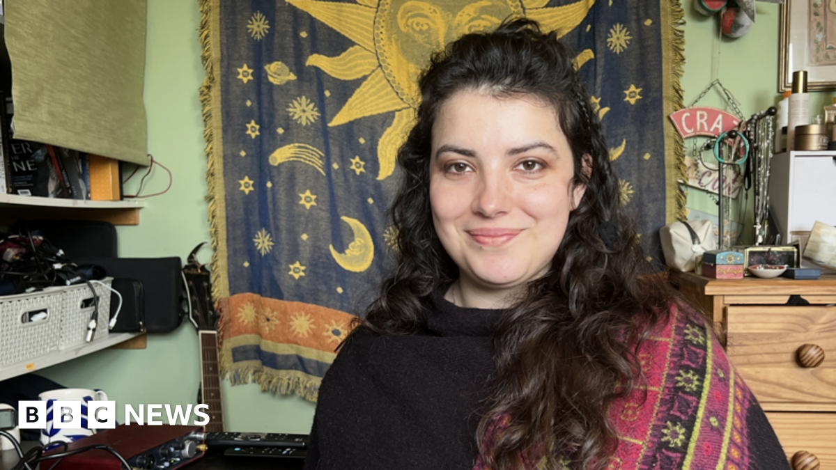 Lily poses for a photograph next to her recording and mixing equipment in her bedroom in Leominster. Behind her are shelves of electronic equipment, a tapestry with the Sun, moons and stars, and a guitar.
