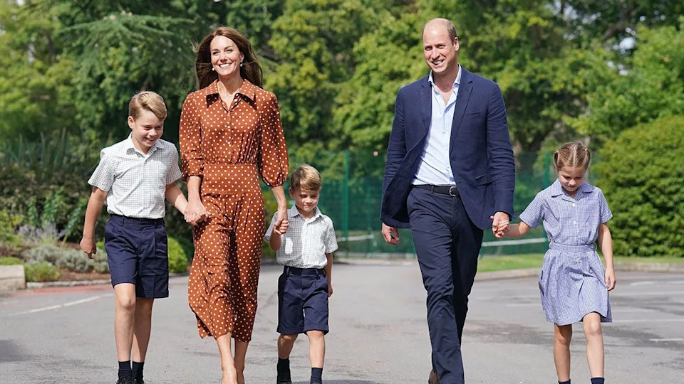 Prince William smiling next to Kate Middleton in a brown polka dot dress as they walk their children to school
