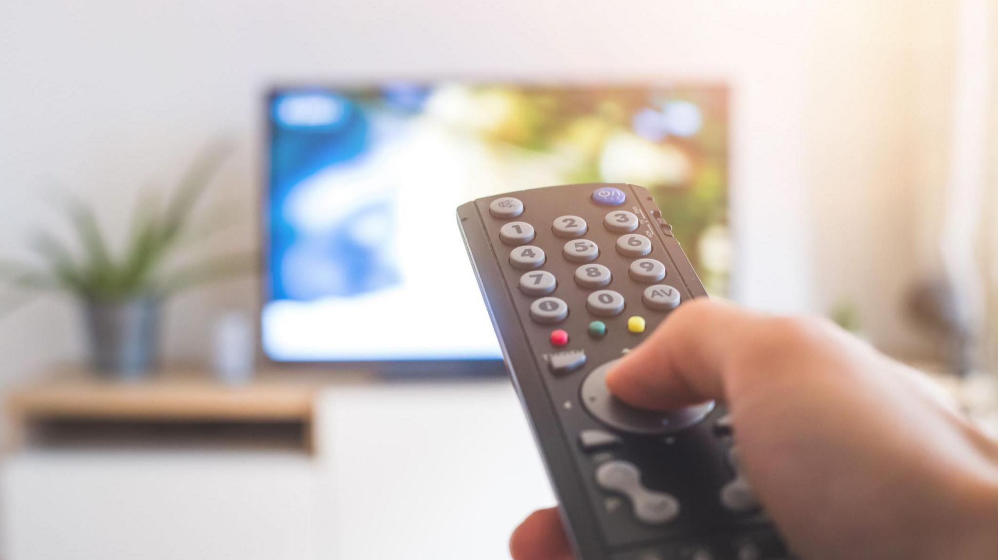 A stock image of someone watching TV. You can see a hand holding a black remote control, the background is out of focus but you can see the outline of a TV screen. 