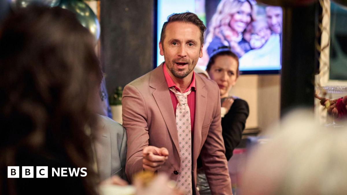 A group of people at a social gathering, Tom Basden is central wearing a pink suit and tie.