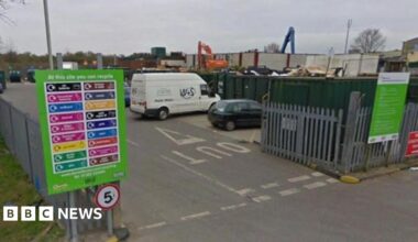 Entrance to a household recycling centre. Cars and vans are lined up in bays in front of open top shipping containers. The site is surrounded by a large grey steel fence.