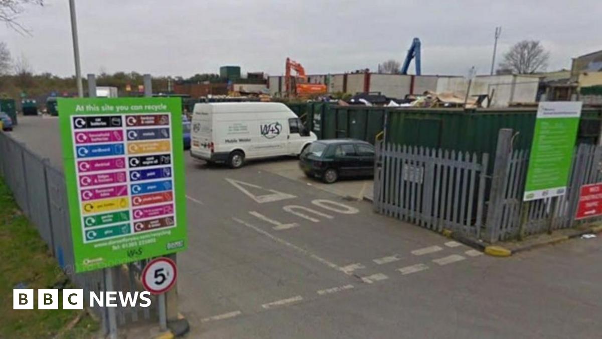 Entrance to a household recycling centre. Cars and vans are lined up in bays in front of open top shipping containers. The site is surrounded by a large grey steel fence.