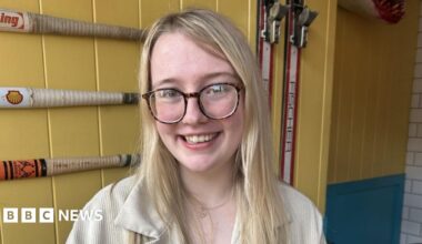 A woman with long blonde hair and brown glasses in sat inside a building with yellow walls. She is smiling at the camera
