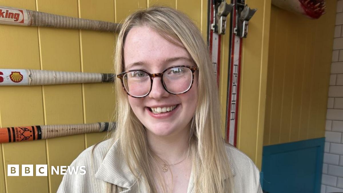 A woman with long blonde hair and brown glasses in sat inside a building with yellow walls. She is smiling at the camera