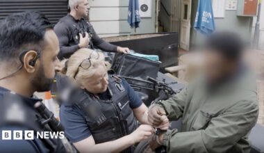 Three immigration officers putting handcuffs on a man. He has dark skin and is wearing a green jacket. There is an e-bike behind him. His face has been blurred so we do not know who he is.