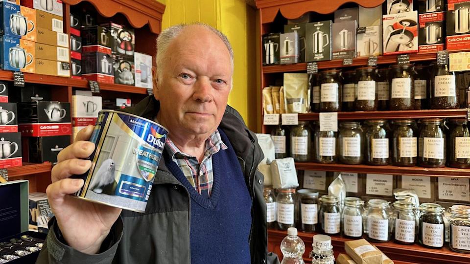John is holding a can of paint to the camera. There are lots of bottles with loose tea in the on shelves in the background.