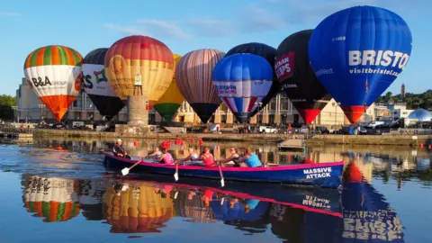 PA Media Several hot air balloons tethered to the ground in Bristol Harbour. There is a small row boat in the foreground of the picture. 