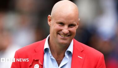 Andrew Strauss with a bald head smiling at the camera. He is wearing a pale blue shirt with a red blazer that has a red rosette on the left.