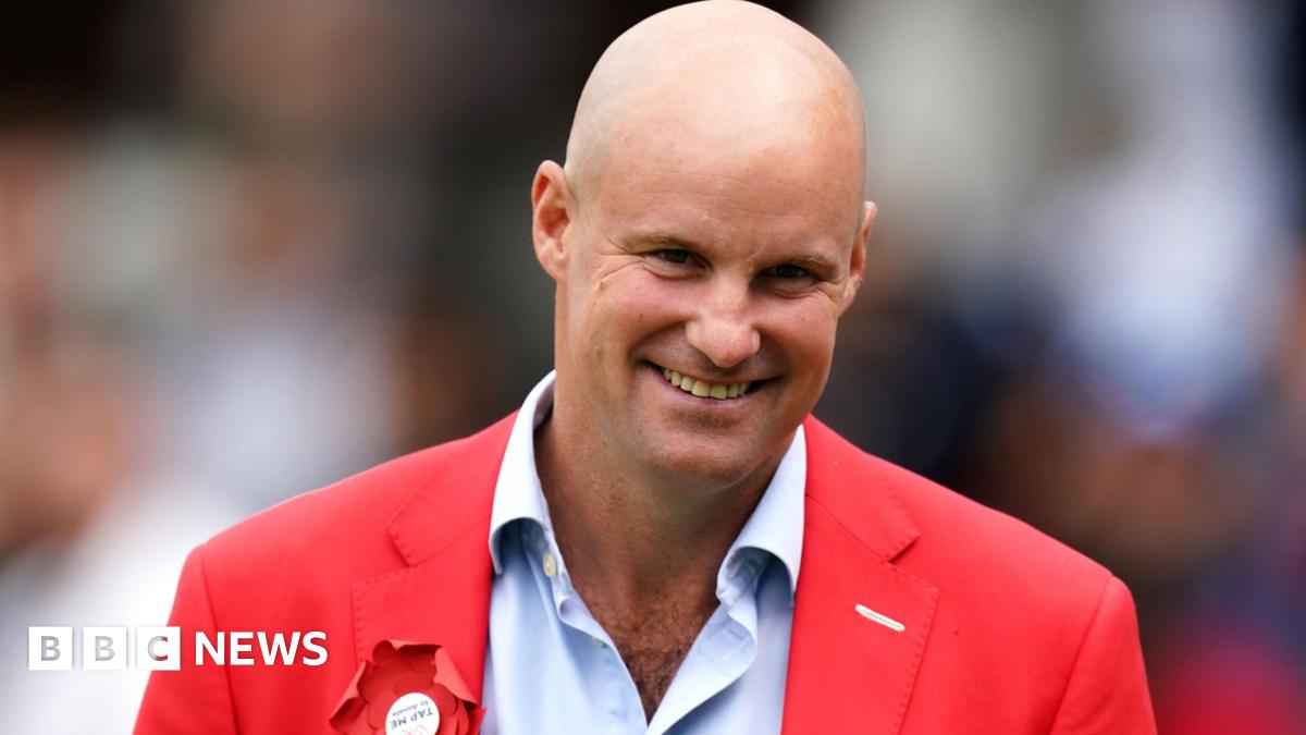 Andrew Strauss with a bald head smiling at the camera. He is wearing a pale blue shirt with a red blazer that has a red rosette on the left.