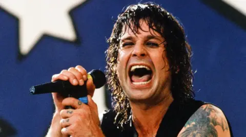 Getty Images A picture of a younger Ozzy Osbourne singing into a microphone, with long brown curly hair and tattoos visible on his arm and fingers, and wearing a black waistcoat and holding a microphone. 