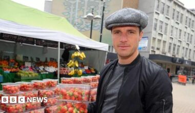 Tommy Coyle wearing a flat cap, standing next to the fruit stall in Hull City Centre. He is holding multiple packs of strawberries and is wearing a black zip up jacket.