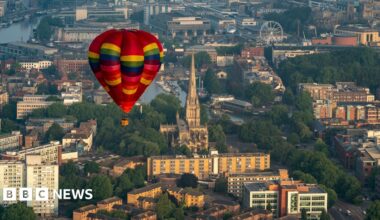 A multi-coloured, mainly red, hot air balloon flying over Bristol. Trees, buildings, and some of the city's landmarks can be seen beneath it including Bristol Cathedral, the harbourside and a ferris wheel.