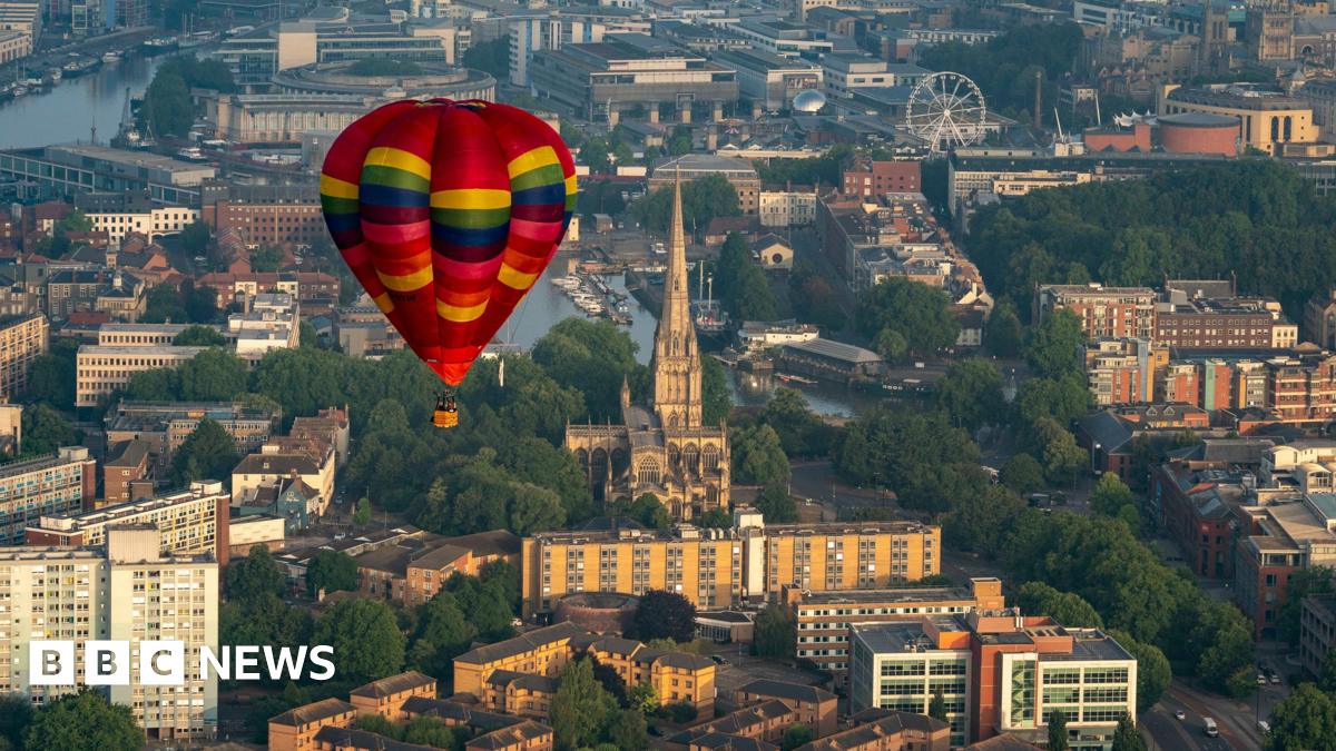 A multi-coloured, mainly red, hot air balloon flying over Bristol. Trees, buildings, and some of the city's landmarks can be seen beneath it including Bristol Cathedral, the harbourside and a ferris wheel.