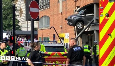 Police officers stand guard next to a cordon, with a car hanging out of the first storey of a large car park in Leeds city centre