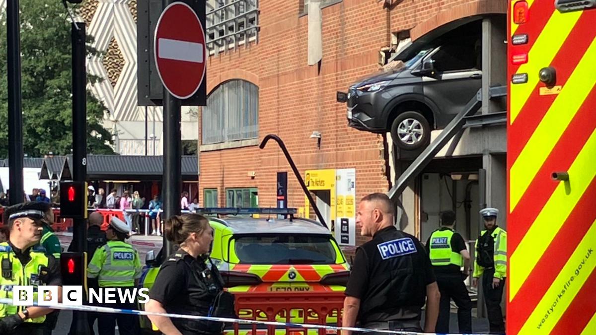 Police officers stand guard next to a cordon, with a car hanging out of the first storey of a large car park in Leeds city centre