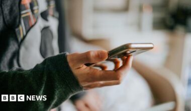 A smartphone held in the hand of an unidentifiable man, who is wearing a colourful top and sitting on a bed.