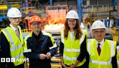A group of three men and a woman stand in what appears to be a manufacturing plant. They all wear hi-vis jackets and helmets.