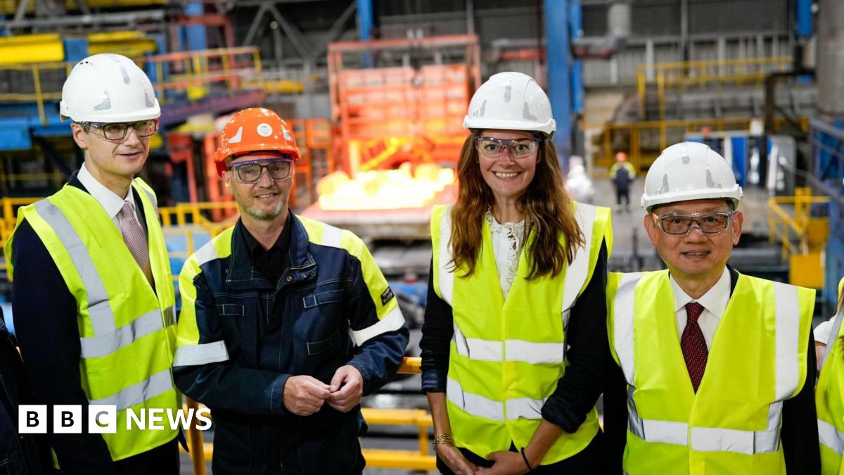 A group of three men and a woman stand in what appears to be a manufacturing plant. They all wear hi-vis jackets and helmets.