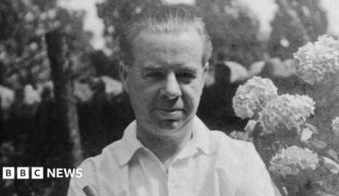 Clement Ernest Kille is seen in black and white wearing a white shirt and standing in front of a flower bush. He has his hair combed back and is smiling.