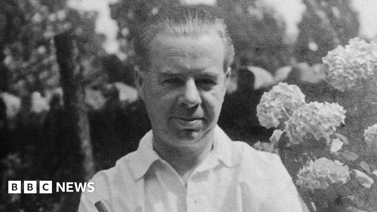 Clement Ernest Kille is seen in black and white wearing a white shirt and standing in front of a flower bush. He has his hair combed back and is smiling.