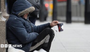 A man in a coat and tracksuit trousers holding up a coffee cup, asking for donations.