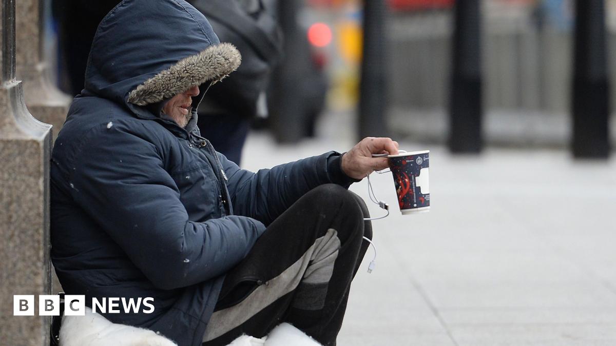 A man in a coat and tracksuit trousers holding up a coffee cup, asking for donations.