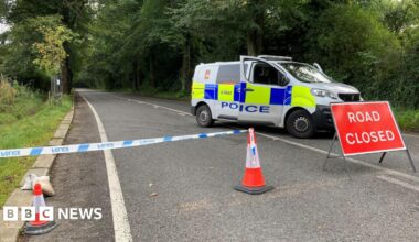 The image shows a police cordon extending to the middle of the road. A van is parked on the right, blocking the way, with a red "Road closed" sign in front of it.