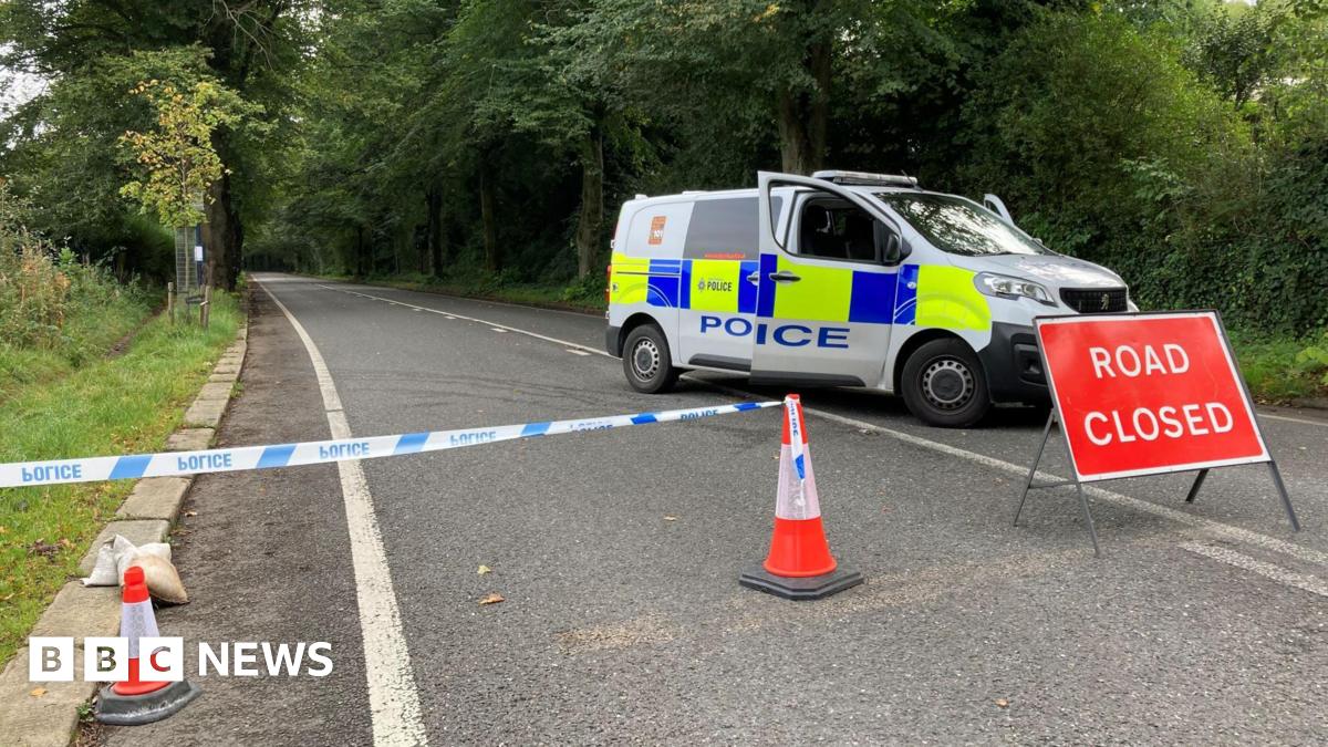 The image shows a police cordon extending to the middle of the road. A van is parked on the right, blocking the way, with a red "Road closed" sign in front of it.