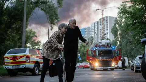 DSNS An elderly woman and man with ambulances and smoke in the sky behind them