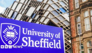 A blue University of Sheffield sign, with the writing in a white font and a university crest to the left. The crest features a book with the writing 'Disce Doce', and is surrounded by arrows, a crown and a white rose. University buildings can be seen behind the sign.