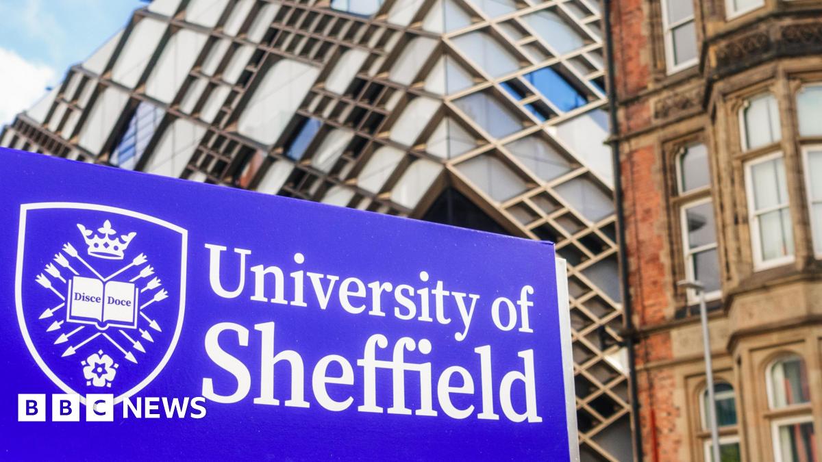 A blue University of Sheffield sign, with the writing in a white font and a university crest to the left. The crest features a book with the writing 'Disce Doce', and is surrounded by arrows, a crown and a white rose. University buildings can be seen behind the sign.