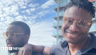 A close-up of two people wearing matching orange-tinted sunglasses. The man on the right is smiling and has his arm on the shoulder of the man on the left, who looks happy but is looking away from the camera. Clouds are in the blue sky and a block of flats is in the background.