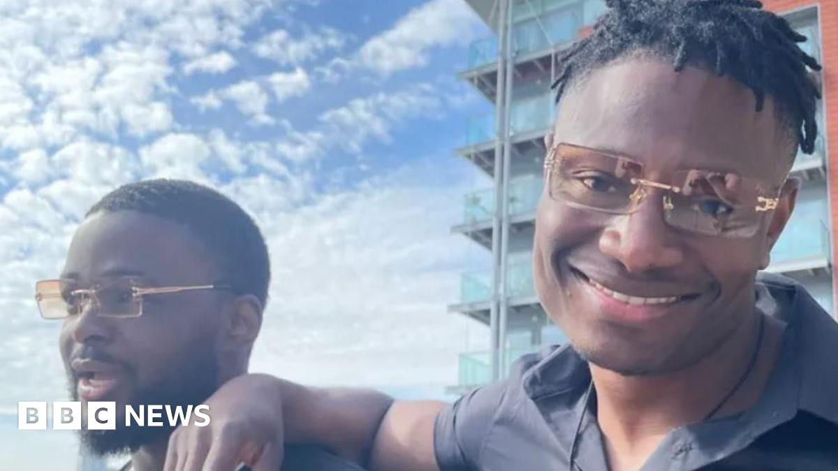 A close-up of two people wearing matching orange-tinted sunglasses. The man on the right is smiling and has his arm on the shoulder of the man on the left, who looks happy but is looking away from the camera. Clouds are in the blue sky and a block of flats is in the background.