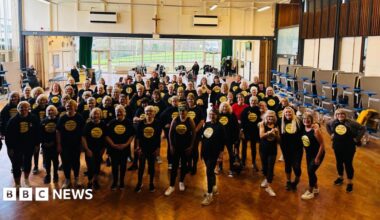 Dozens of middle aged and elderly women stood in a large hall with a wooden floor. They are wearing matching black t-shirts, which have a yellow circle in the centre.
