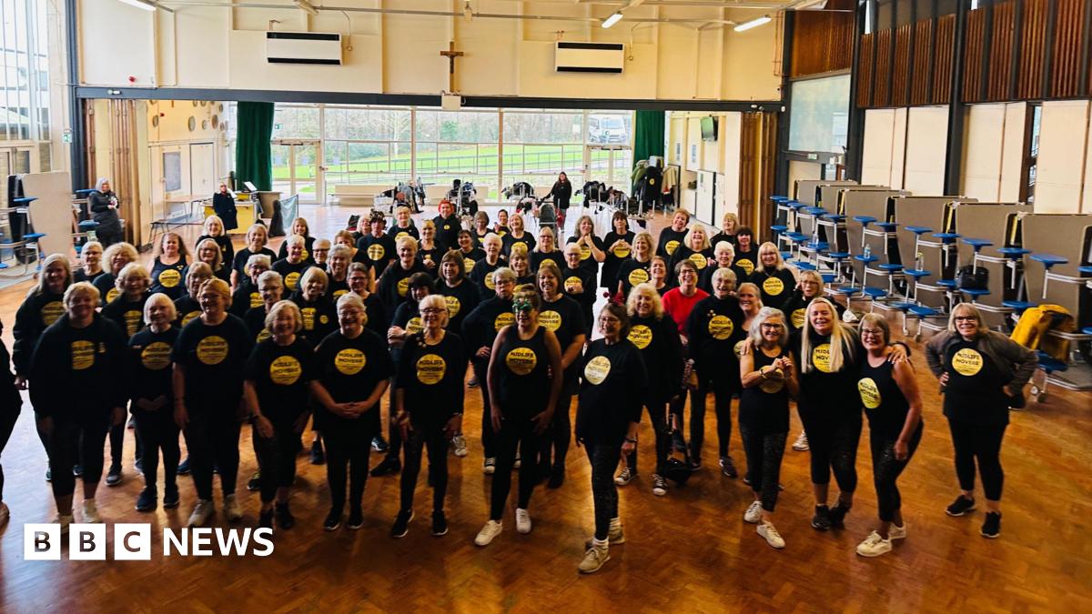Dozens of middle aged and elderly women stood in a large hall with a wooden floor. They are wearing matching black t-shirts, which have a yellow circle in the centre.