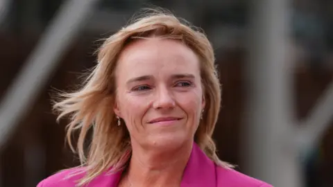 PA Media Sandie Peggie - a woman with blond hair to her shoulders and a pink jacket - smiles while standing outside 