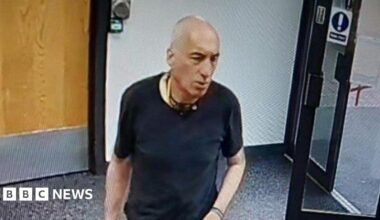 A man in a black t-shirt stood in a room with wooden internal door and an external glass door, which is open. There is a plain wall behind him and a carpeted floor.