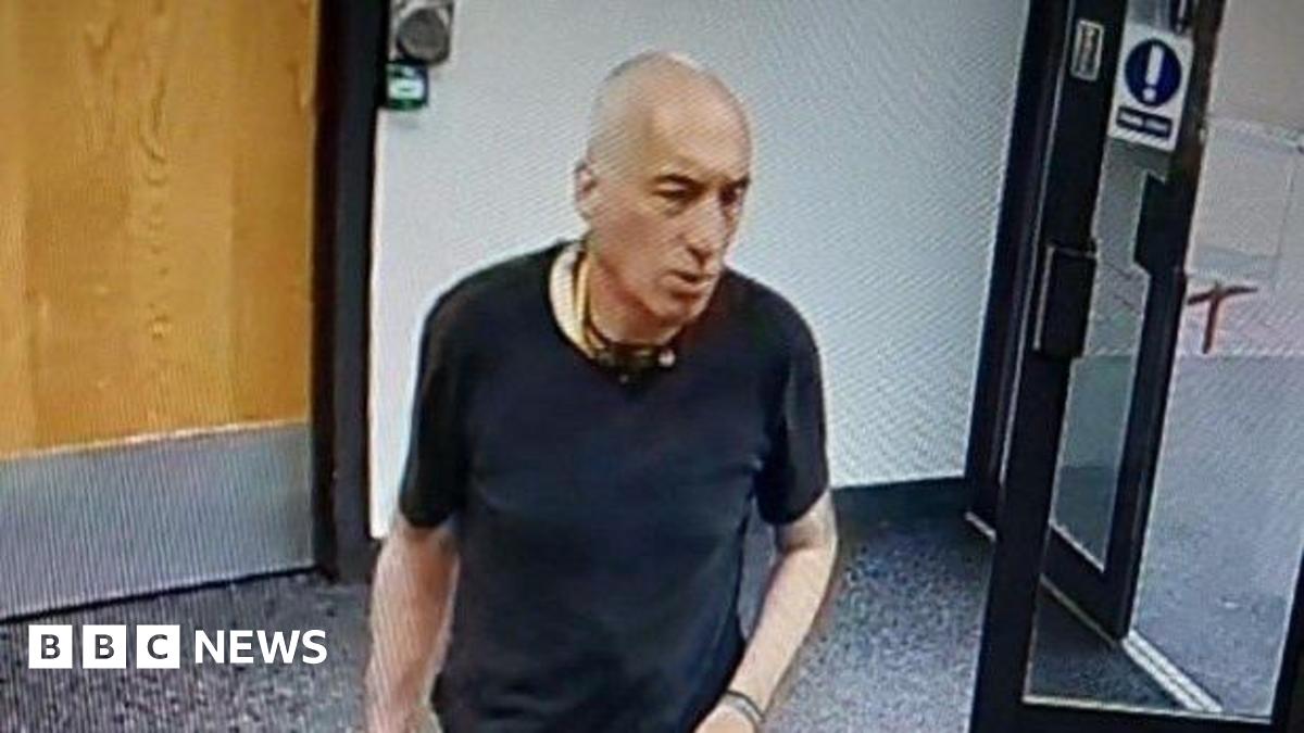 A man in a black t-shirt stood in a room with wooden internal door and an external glass door, which is open. There is a plain wall behind him and a carpeted floor.
