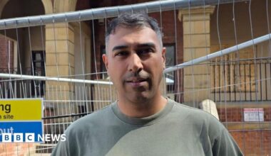 A man in a green t-shirt stands in front of railings next to a large old brick building