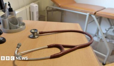 A brown-red stethoscope on a desk in a doctor's surgery. A medical bed is in the background.