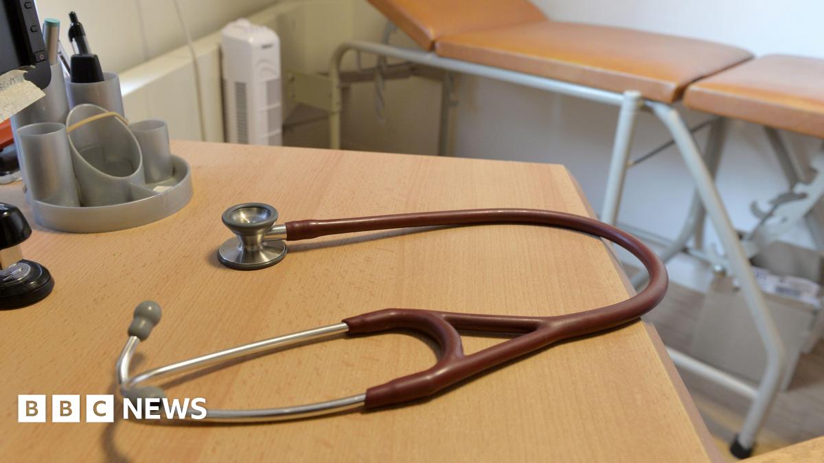 A brown-red stethoscope on a desk in a doctor's surgery. A medical bed is in the background.
