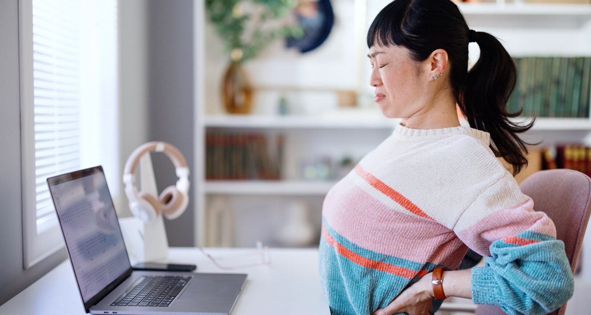 woman wearing a striped jumper sitting at a desk in front of a laptop, holding her lower back and grimacing, sideways onto the camera