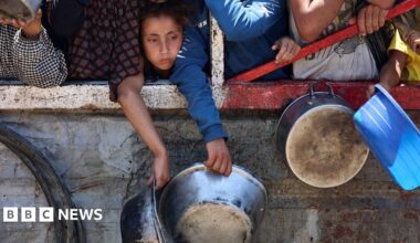 A young Palestinian girl hangs over a wall, holding a metal bowl, while waiting for lentil soup distribution point in Gaza City in the northern Gaza Strip