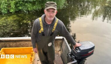 A middle aged white man is on a boat on a lake. He wears a grey hoodie, yellow straps and waterproof green dungarees. There is a yellow crate on the boat.