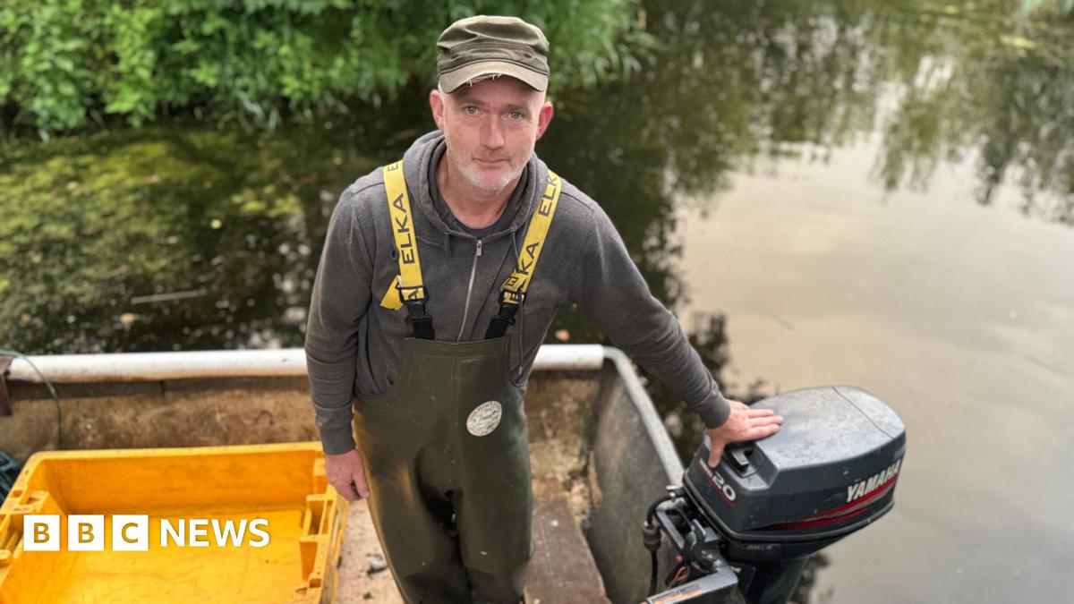 A middle aged white man is on a boat on a lake. He wears a grey hoodie, yellow straps and waterproof green dungarees. There is a yellow crate on the boat.